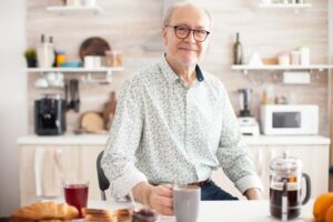 Senior man in kitchen smiling looking at camera holding hot coffee cup.